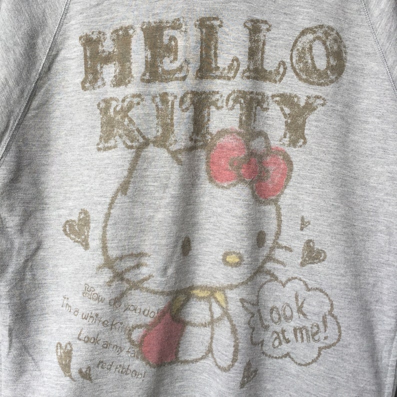 Op de afbeelding: Grijs sweatshirt met een Hello Kitty-print. De print toont Hello Kitty die een rood hart vasthoudt en een spraakballon met de tekst "Kijk naar me!" De tekst "How do you do I'm a white Kitty Look at my face red ribbon!" staat onder de print.