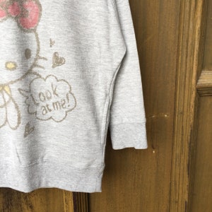 Op de afbeelding: Grijze longsleeve trui met een Hello Kitty-print en de tekst "look at me!" in een spraakballon.