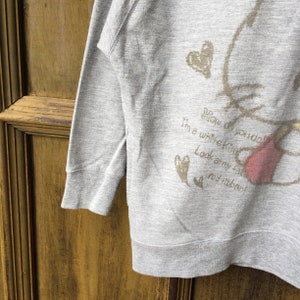 Op de afbeelding: Grijs sweatshirt met een cartoonillustratie van een witte kat met een rode strik. De tekst op het sweatshirt luidt "How do you do! I'm a white kitty. Look at my big red ribbon!"