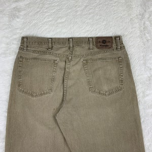 Op de afbeelding: Een paar bruine Wrangler jeans met een bruin leren patch op de achterzak met de tekst "Wrangler" in wit.