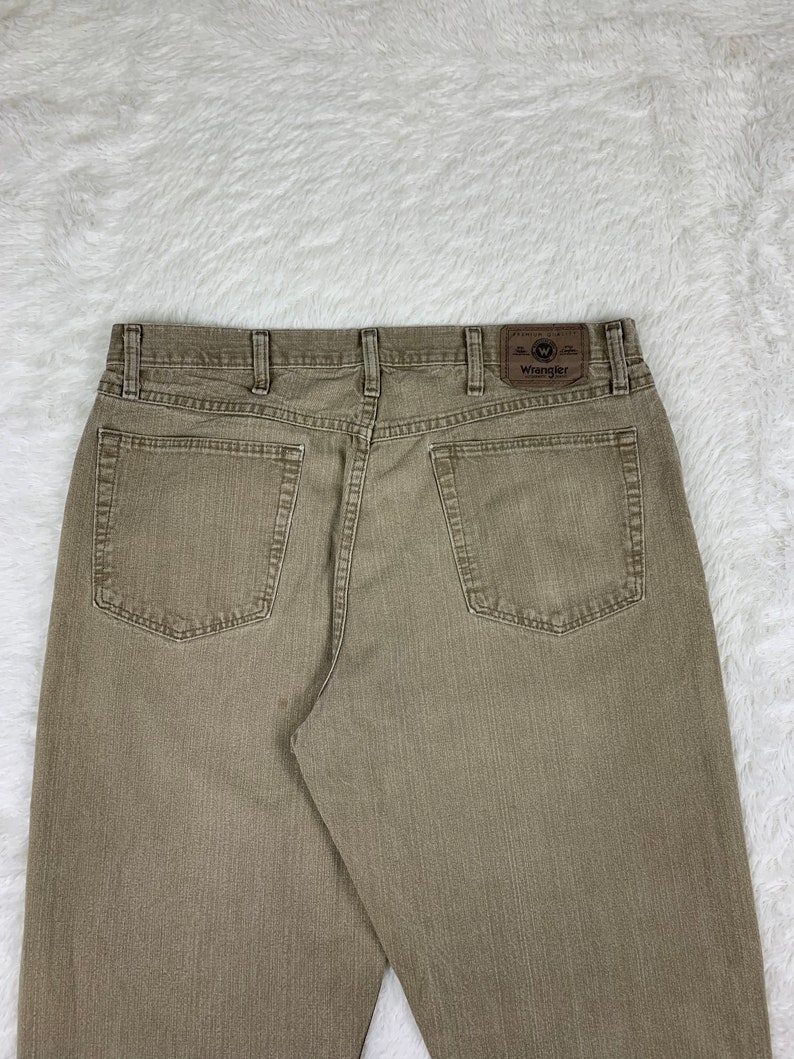 Op de afbeelding: Een paar bruine Wrangler jeans met een bruin leren patch op de achterzak met de tekst "Wrangler" in wit.