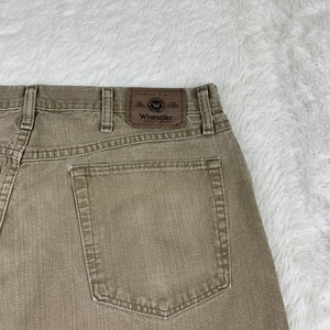 Op de afbeelding: Een paar bruine Wrangler jeans met een achterzak en een bruin leren patch met het Wrangler-logo en de tekst "Premium Quality" en "Authentic Jeans".