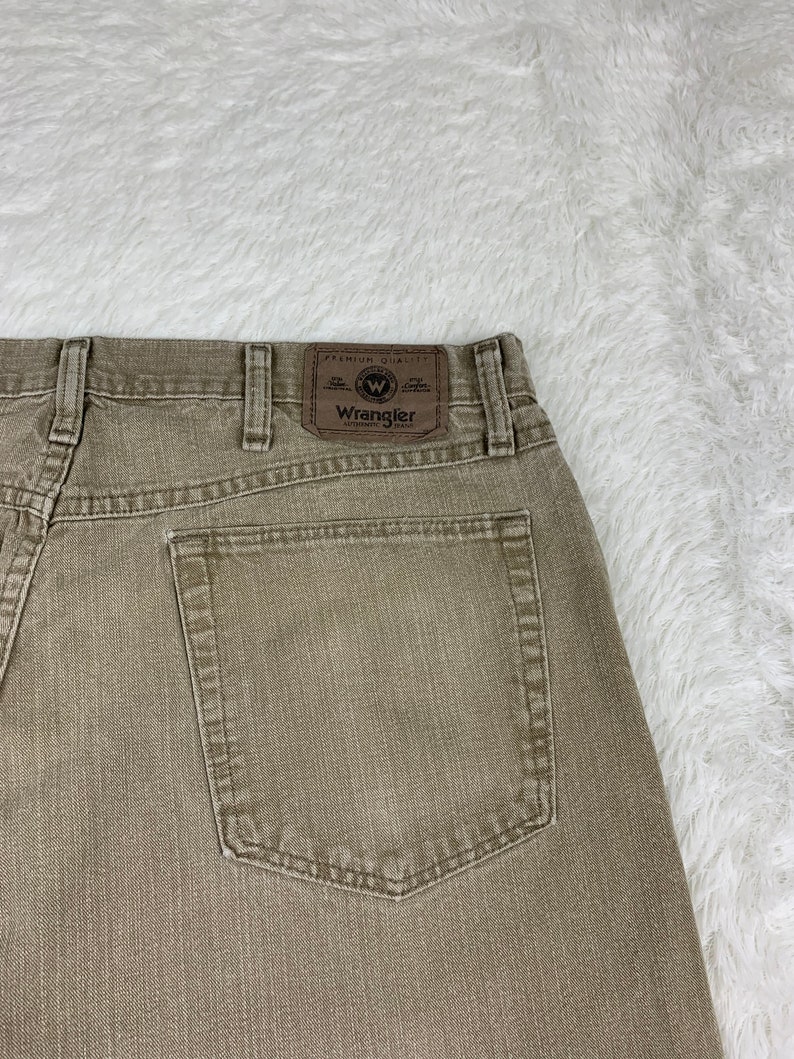 Op de afbeelding: Een paar bruine Wrangler jeans met een achterzak en een bruin leren patch met het Wrangler-logo en de tekst "Premium Quality" en "Authentic Jeans".