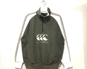 Puede incluir: Sudadera con cremallera Canterbury verde oliva con mangas blancas y rayas doradas. La sudadera presenta un cuello alto, una cremallera negra y el logotipo de Canterbury bordado en dorado.