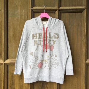 Op de afbeelding: Grijze hoodie met een Hello Kitty-print. De print toont Hello Kitty in een roze jurk met een rode strik, die een rood hart vasthoudt. De tekst "Hello Kitty" is in bruin boven de print gedrukt. De hoodie heeft een rood trekkoord.