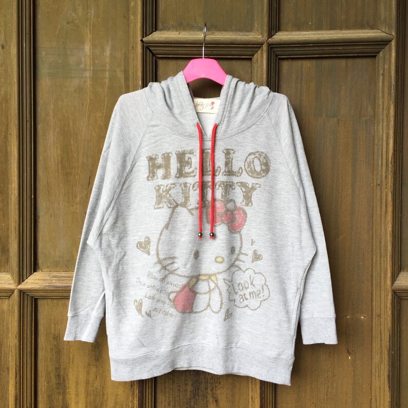 Op de afbeelding: Grijze hoodie met een Hello Kitty-print. De print toont Hello Kitty in een roze jurk met een rode strik, die een rood hart vasthoudt. De tekst "Hello Kitty" is in bruin boven de print gedrukt. De hoodie heeft een rood trekkoord.
