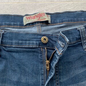 K&ouml;nnte beinhalten: Eine blaue Jeanshose mit Rei&szlig;verschluss und einem Wrangler-Markenlabel. Die Jeans hat einen Knopfverschluss und einen Metallknopf mit dem Wrangler-Logo.