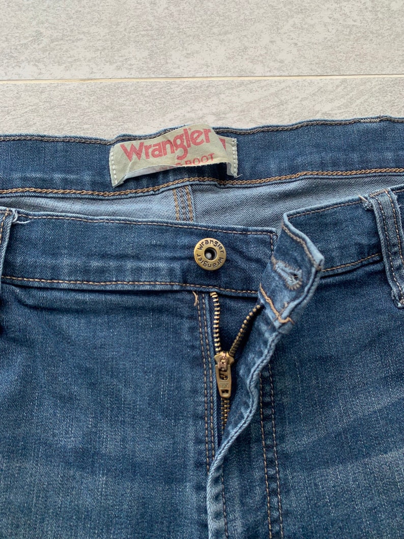 K&ouml;nnte beinhalten: Eine blaue Jeanshose mit Rei&szlig;verschluss und einem Wrangler-Markenlabel. Die Jeans hat einen Knopfverschluss und einen Metallknopf mit dem Wrangler-Logo.