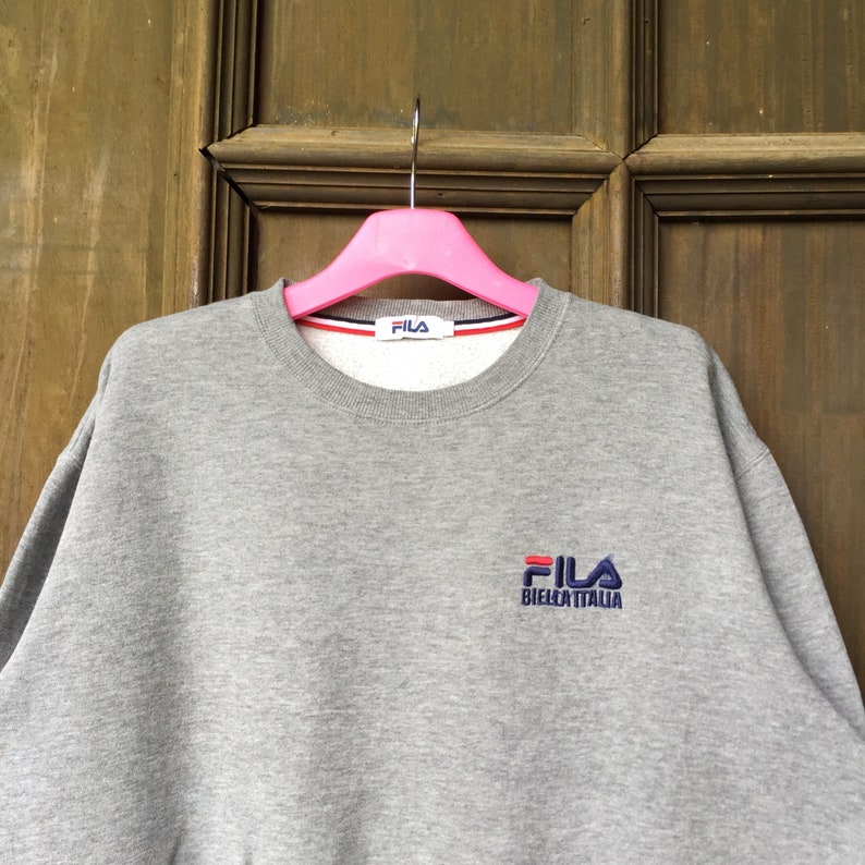 May include: Gray sweatshirt with a Fila logo embroidered on the front. The logo is in red, blue, and white and says "Fila Biella Italia".