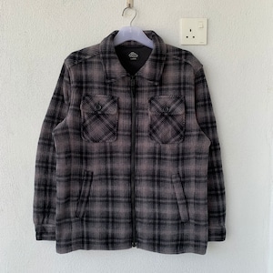 Può includere: Una camicia di flanella a quadri neri e grigi con chiusura a zip. La camicia ha due tasche a patta sul petto e un'etichetta con la scritta "Thrasher" sul lato sinistro.