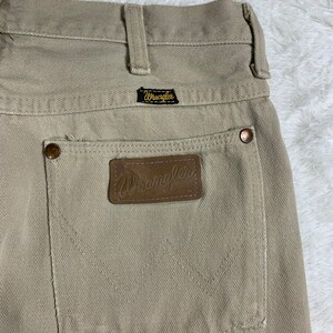 Op de afbeelding: Een beige Wrangler-jeans met een bruine leren patch op de achterzak. De patch heeft het Wrangler-logo in goud gegraveerd.