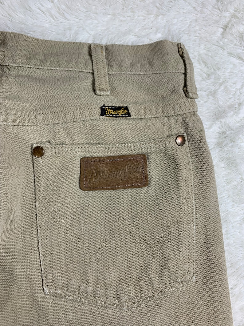 Op de afbeelding: Een beige Wrangler-jeans met een bruine leren patch op de achterzak. De patch heeft het Wrangler-logo in goud gegraveerd.
