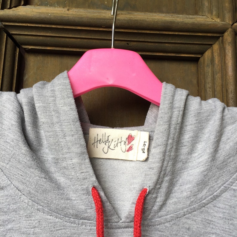 Op de afbeelding: Grijze hoodie met rode trekkoorden en een Hello Kitty-label met twee rode harten. Het label zegt "Hello Kitty" en "Large".