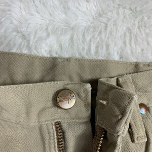 Op de afbeelding: Close-up van de voorzijde van de tailleband van een kaki broek. De broek heeft een ritsvlieg en een knoopsluiting. De knoop heeft het woord "Wrangler" erop gegraveerd.