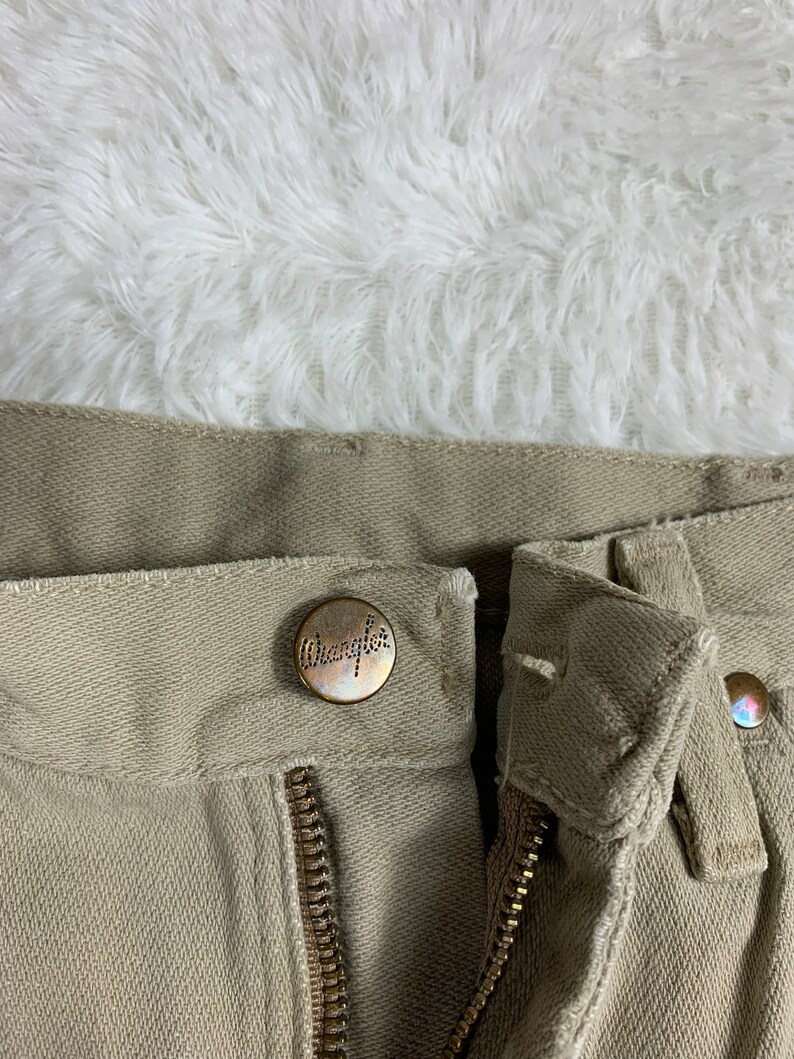 Op de afbeelding: Close-up van de voorzijde van de tailleband van een kaki broek. De broek heeft een ritsvlieg en een knoopsluiting. De knoop heeft het woord "Wrangler" erop gegraveerd.