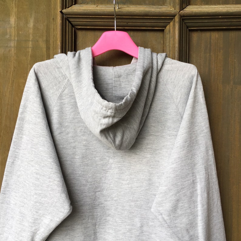 Op de afbeelding: Een grijs hoodie-vest dat aan een roze hanger hangt. De trui heeft een capuchon met trekkoord en lange mouwen.