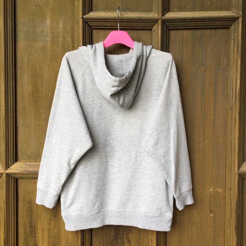 Op de afbeelding: Een grijs hoodie met een capuchon met trekkoord. De hoodie hangt aan een roze hanger tegen een houten achtergrond.