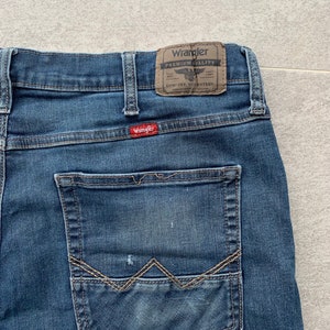 K&ouml;nnte beinhalten: Eine blaue Jeanshose mit einer Ges&auml;&szlig;tasche, die mit einem markanten Steppdesign verziert ist. Die Jeans hat einen braunen Leder-Patch mit dem Text "Wrangler Premium Quality" und einem kleinen Adler-Logo.