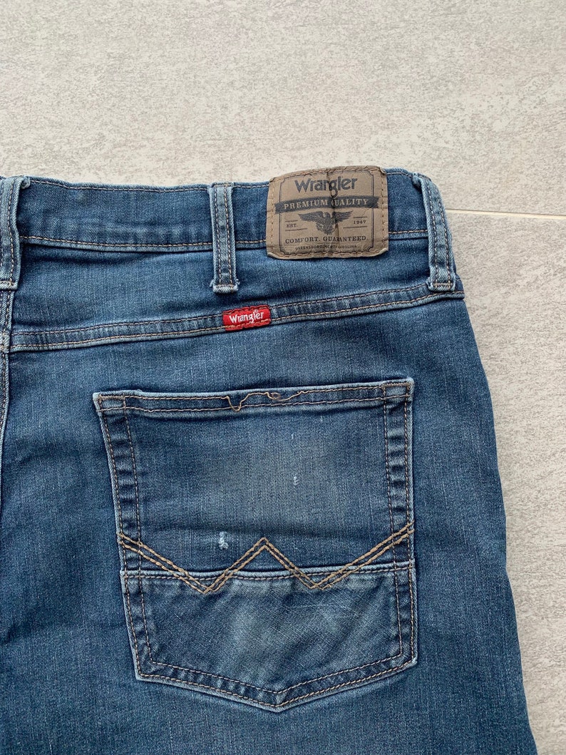 K&ouml;nnte beinhalten: Eine blaue Jeanshose mit einer Ges&auml;&szlig;tasche, die mit einem markanten Steppdesign verziert ist. Die Jeans hat einen braunen Leder-Patch mit dem Text "Wrangler Premium Quality" und einem kleinen Adler-Logo.