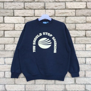 Könnte beinhalten: Marineblaues Sweatshirt mit dem weißen Text "YOU SHOULD STOP SMOKING", der sich um einen hellgelben Kreis mit horizontalen Linien wölbt. Das Sweatshirt hängt an einem hellblauen Kleiderbügel vor einer Backsteinmauer.