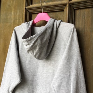 Op de afbeelding: Een grijs hoodie-vest dat aan een roze hanger hangt. De trui heeft een capuchon met trekkoord en lange mouwen.