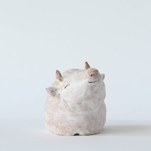 May include: A small, off-white ceramic cow figurine with a smiling expression. The cow has small, brown horns and ears, and its body is rounded. The figurine is painted with a textured, matte finish, and is set against a plain white background.