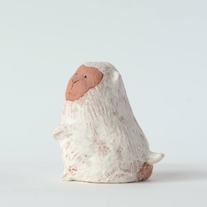 Pode incluir: Pequena figura de cerâmica feita à mão de um animal estilizado. A figura tem um corpo branco e texturizado e um rosto castanho-avermelhado. O animal está sentado com os braços ao lado. A figura tem um design simples e minimalista.