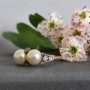 May include: A vintage ring featuring two large, creamy pearls and small diamonds. The ring has a gold band and a silver setting for the diamonds. Pink and white flowers and green leaves are in the background.