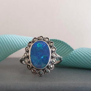 Op de afbeelding: Een zilveren ring met een ovale, iriserende blauwe edelsteen. De edelsteen is gezet in een decoratieve zilveren setting met kleine, donkere accenten. De ring wordt gepresenteerd tegen een lichtblauw lint.