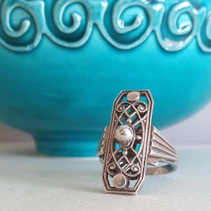 Op de afbeelding: Zilverkleurige filigraanring met een Art Deco-ontwerp. De ring heeft een langwerpige rechthoekige voorkant met ingewikkeld krulwerk, een centrale bol en twee kleinere cirkelvormige accenten. De band van de ring heeft een geribbelde textuur. Op de achtergrond een turquoise keramische kom.