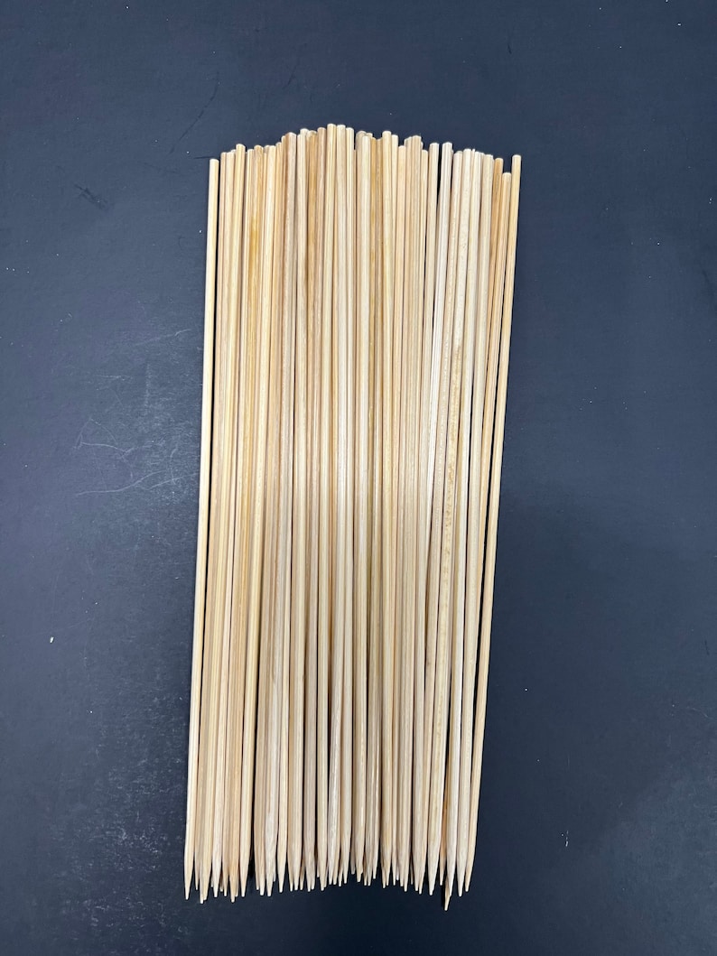 Op de afbeelding: Een bundel natuurlijke houten spiesjes, ongeveer 30 in totaal, met puntige uiteinden. De spiesjes zijn netjes in een rij gerangschikt tegen een donkere achtergrond.