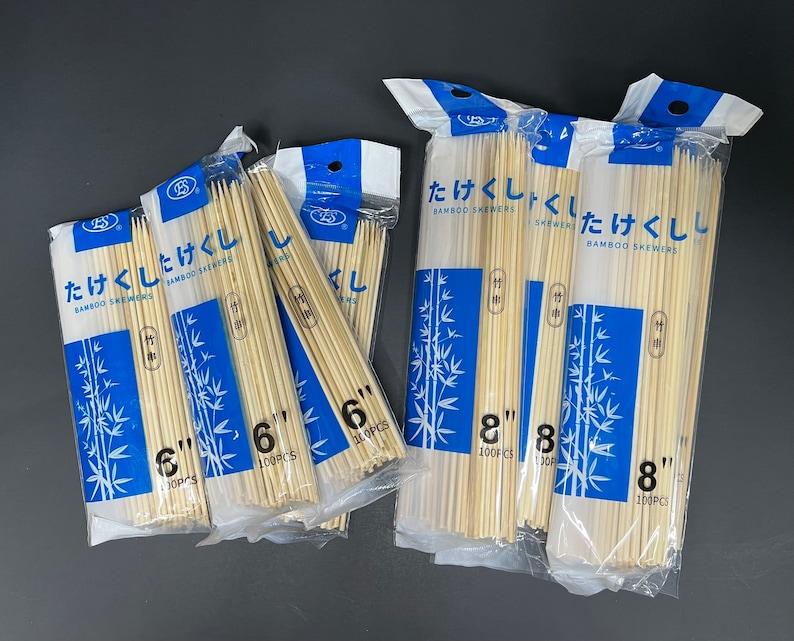 Op de afbeelding: Vier pakketten bamboesprieten, elk met een blauw-wit ontwerp. De pakketten zijn gelabeld "Takekushi Bamboo Skewers" en tonen de lengte van de sprieten in inches. De pakketten zijn in een rij gerangschikt, met het grootste pakket rechts en het kleinste pakket links.