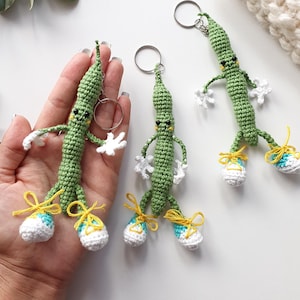 May include: Three green crochet bean keychains with small black eyes, white hands, and blue and white sneakers with yellow laces. Each bean figure has a metal keyring attached to the top, featuring a slender body and articulated limbs.