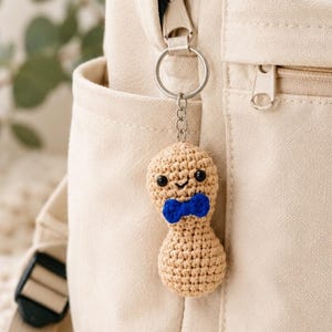 May include: A beige crocheted peanut keychain featuring a small blue bow tie, black bead eyes, and a stitched smile. The peanut is attached to a silver-toned metal split ring and chain, which is clipped onto the zipper of a beige canvas backpack.