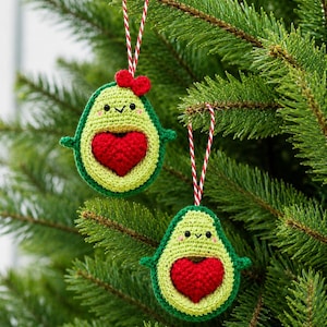 May include: Two crocheted avocado ornaments featuring light green flesh, dark green skin, and a central red heart-shaped pit. Each avocado has small black eyes, a stitched smile, and is suspended by a red and white twisted string loop.