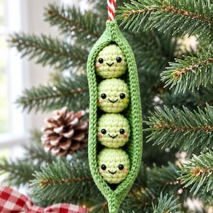 May include: A crocheted green pea pod ornament featuring four small, round, light green peas with embroidered black eyes, smiling mouths, and pink cheeks. The pod is suspended by a red and white striped string for hanging on a tree branch.