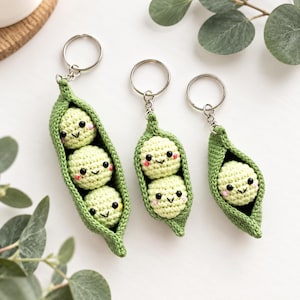 May include: Three crochet keychains featuring green pea pods with smiling faces. One pod contains three peas, one contains two peas, and one contains a single pea. Each pea has black bead eyes, a stitched mouth, and pink cheeks.