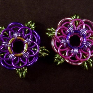 May include: Two purple and pink flower-shaped jewelry pieces with gold accents. The flowers are made of metal rings and have a green leaf design.