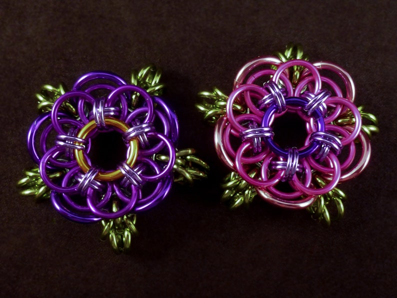 May include: Two purple and pink flower-shaped jewelry pieces with gold accents. The flowers are made of metal rings and have a green leaf design.