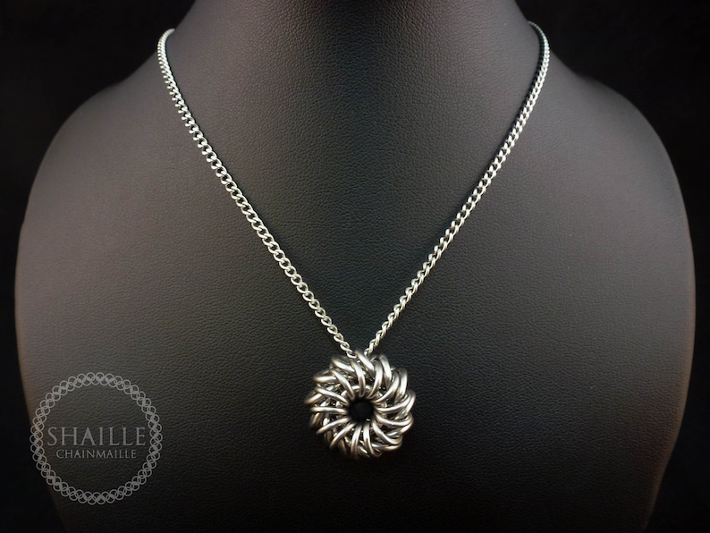 May include: A silver chain necklace with a silver chainmaille pendant in the shape of a circle. The pendant has a small hole in the center.