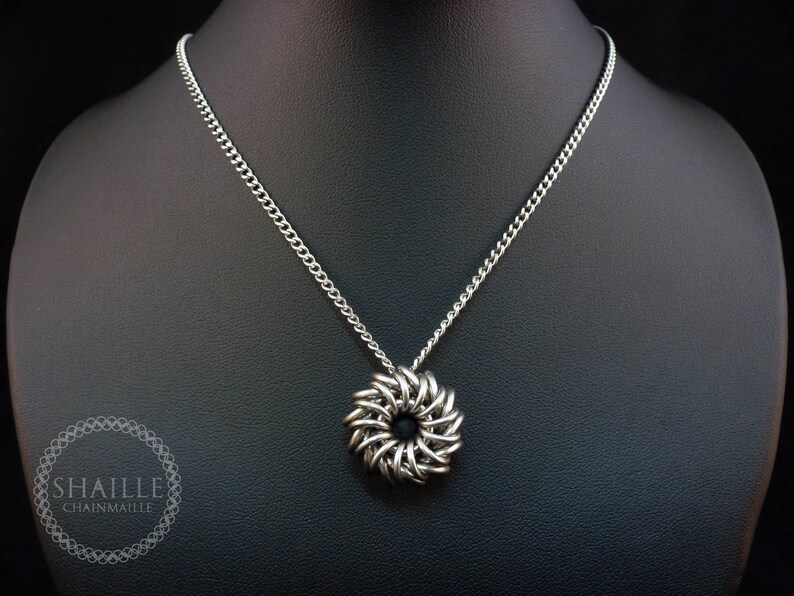 May include: A silver chain necklace with a silver chainmaille pendant in the shape of a flower. The pendant has a black center.