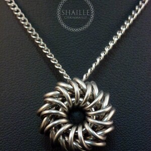 May include: A silver chainmail necklace with a circular pendant. The pendant is made of interwoven metal rings and has a dark center.