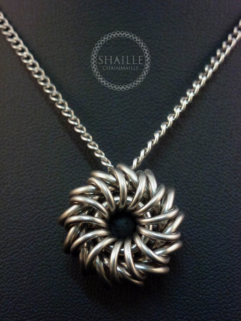 May include: A silver chainmail necklace with a circular pendant. The pendant is made of interwoven metal rings and has a dark center.