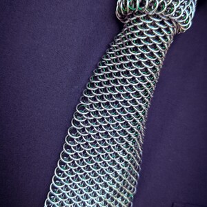 Puede incluir: Una corbata de cota de malla plateada con interior verde, sobre un fondo morado oscuro. La corbata est&aacute; hecha de anillos met&aacute;licos entrelazados, mostrando un dise&ntilde;o detallado y texturizado. El logotipo "SHAILE CHAINMAILLE" es visible.