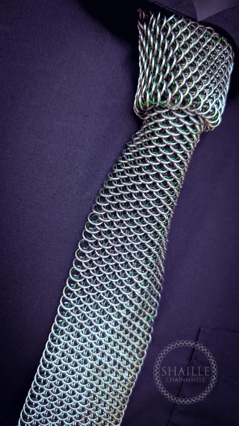 Puede incluir: Una corbata de cota de malla plateada con interior verde, sobre un fondo morado oscuro. La corbata est&aacute; hecha de anillos met&aacute;licos entrelazados, mostrando un dise&ntilde;o detallado y texturizado. El logotipo "SHAILE CHAINMAILLE" es visible.