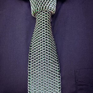 Puede incluir: Una corbata de cota de malla plateada con detalles verdes, sobre una camisa azul oscuro. La corbata est&aacute; hecha de anillos met&aacute;licos entrelazados, creando un patr&oacute;n texturizado en forma de escamas. La marca "SHAILLE CHAINMAILLE" es visible.