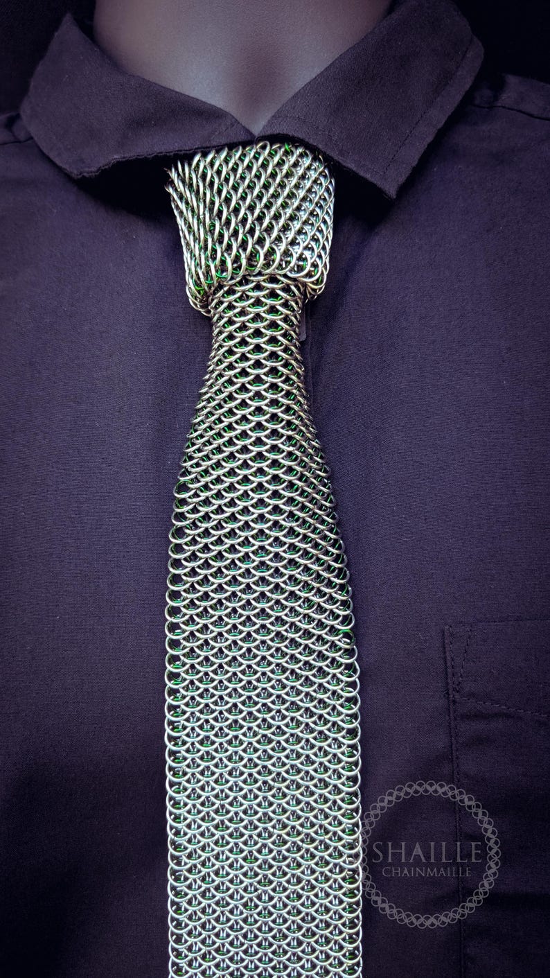Puede incluir: Una corbata de cota de malla plateada con detalles verdes, sobre una camisa azul oscuro. La corbata est&aacute; hecha de anillos met&aacute;licos entrelazados, creando un patr&oacute;n texturizado en forma de escamas. La marca "SHAILLE CHAINMAILLE" es visible.