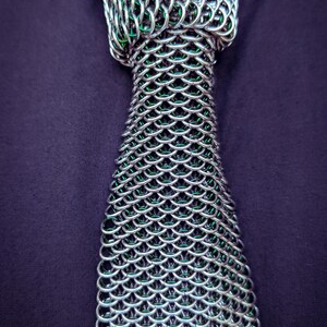 Puede incluir: Una corbata de cota de malla plateada con interior verde, exhibida sobre una camisa morada oscura. La corbata est&aacute; construida con anillos de metal entrelazados, creando un patr&oacute;n texturizado en forma de escamas. El dise&ntilde;o de la corbata es visible desde el nudo.
