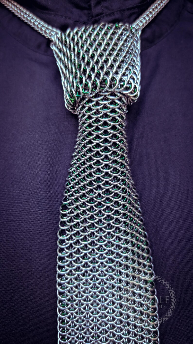 Puede incluir: Una corbata de cota de malla plateada con interior verde, exhibida sobre una camisa morada oscura. La corbata est&aacute; construida con anillos de metal entrelazados, creando un patr&oacute;n texturizado en forma de escamas. El dise&ntilde;o de la corbata es visible desde el nudo.