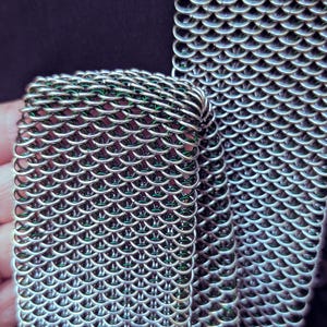Puede incluir: Primer plano de una secci&oacute;n de cota de malla. Los eslabones met&aacute;licos est&aacute;n intrincadamente entrelazados, creando un patr&oacute;n flexible en forma de escamas. La cota de malla es de color plateado, con un ligero brillo, sobre un fondo oscuro.